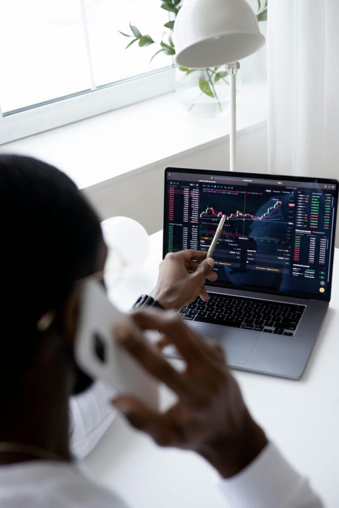 A businessman using a smartphone while analysing financial trading graphs on a laptop indoors.